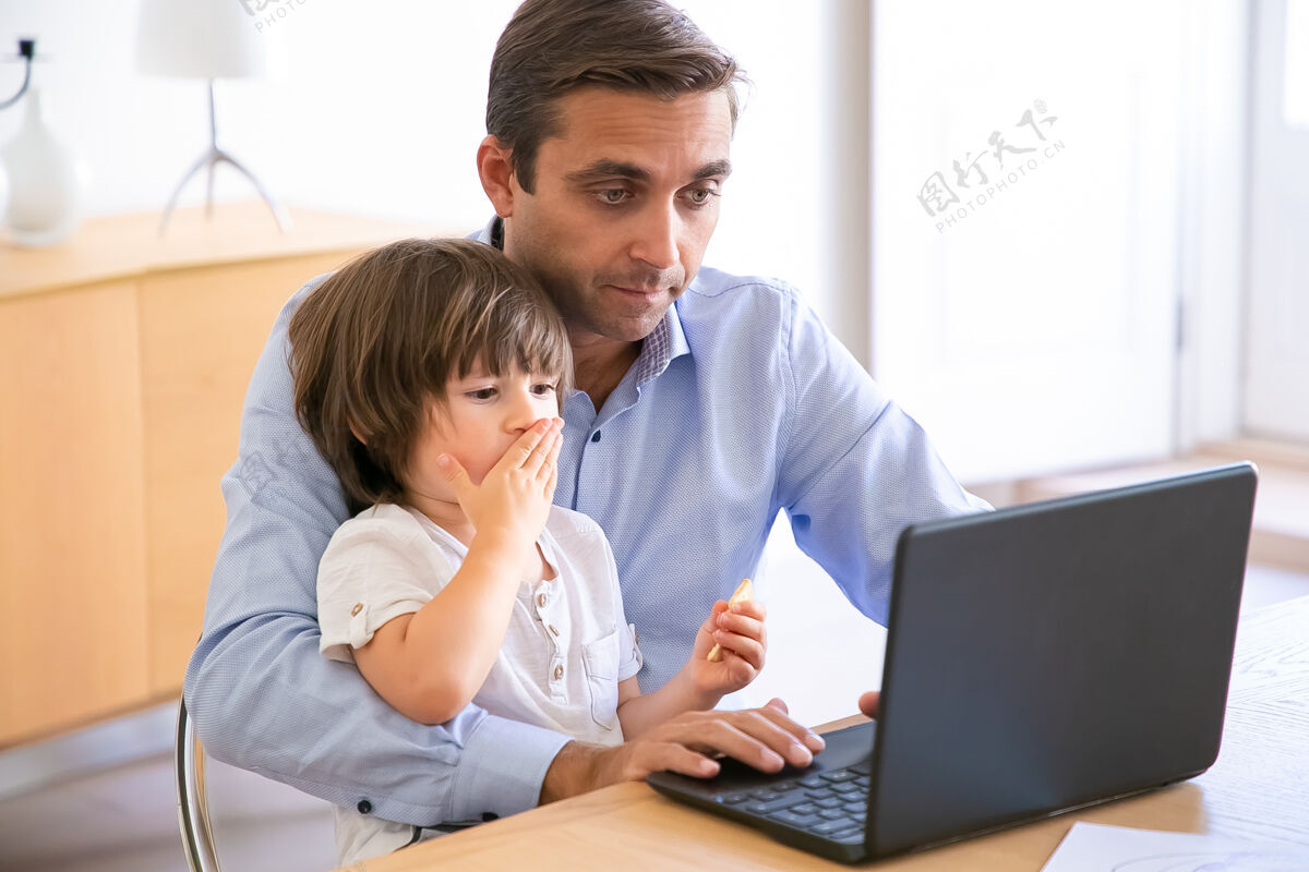 集中专注的爸爸用笔记本电脑抱着儿子跪着高加索中年爸爸和可爱的小男孩坐在桌子旁 用电脑工作童年 自由职业者和父亲的概念自由职业者父亲玩耍