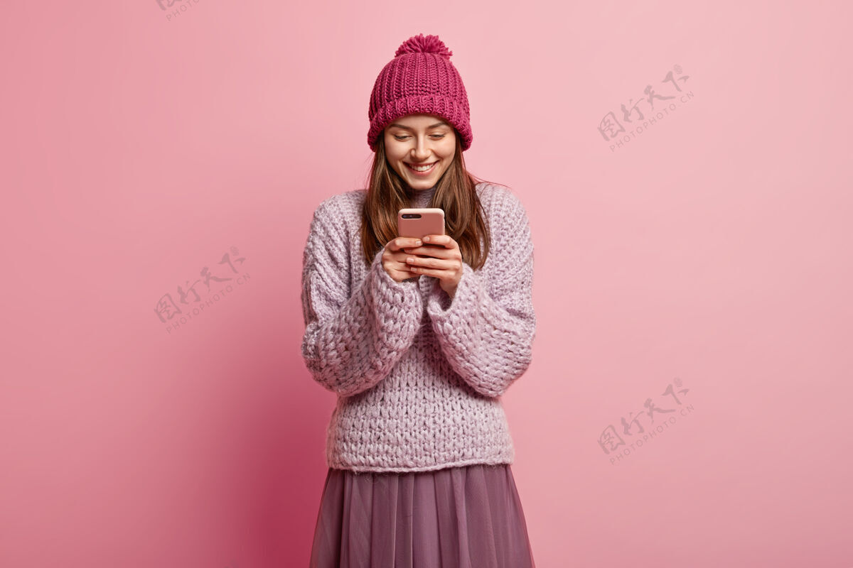手机快乐乐观的年轻女子手持智能手机 戴着冬季帽子和针织套头衫毛衣手机小工具
