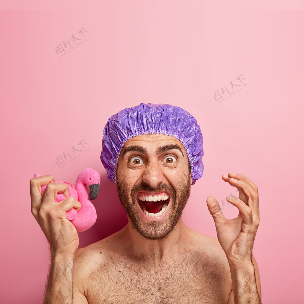 男情绪激动的男人积极地打手势 恼火地拿着柔软的粉红色火烈鸟海绵 戴着浴帽凝视身体护理男性