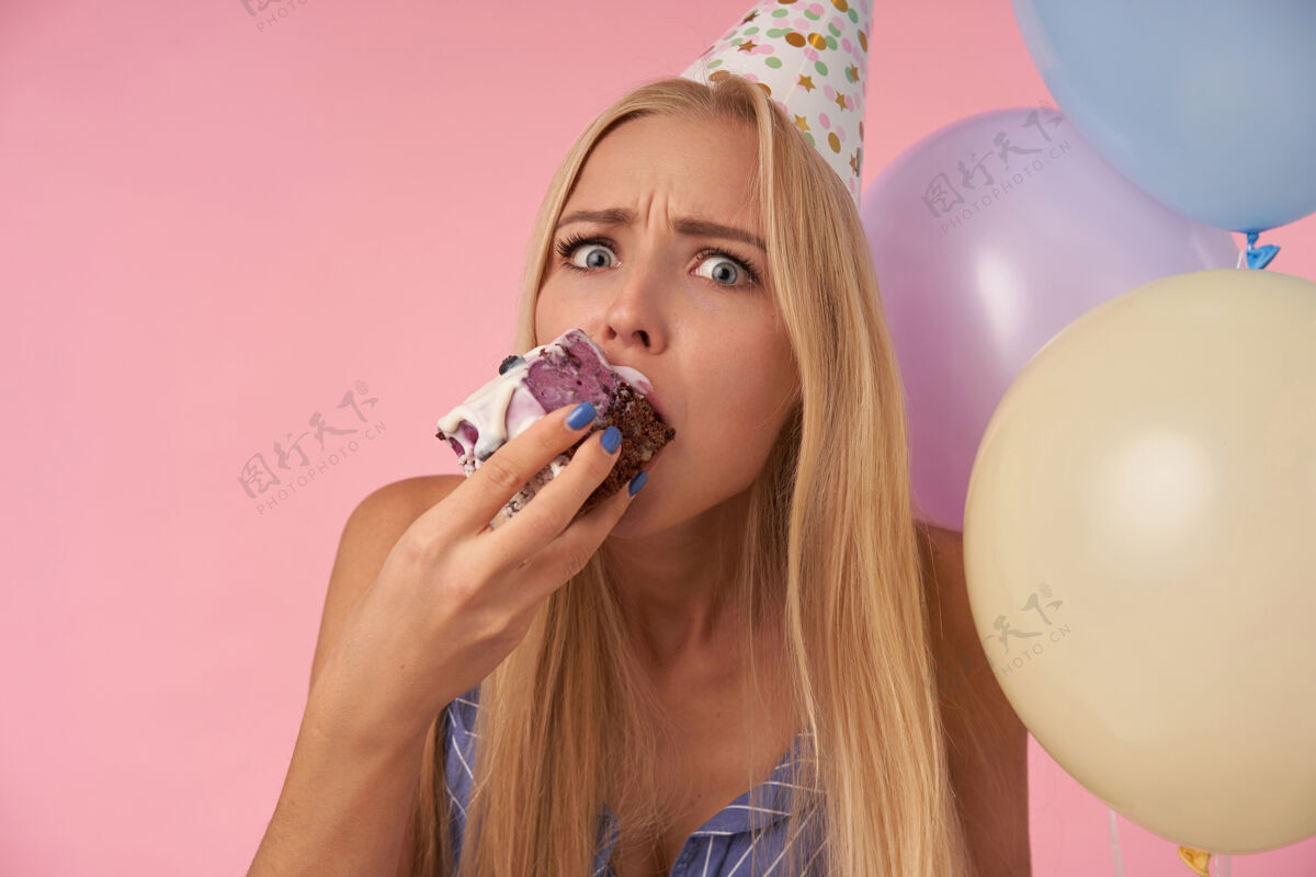 气球特写镜头中困惑的年轻长发金发女性戴着节日锥形帽 在五颜六色的气球上吃着生日蛋糕 看着相机 眉头紧锁金发饮食皱眉