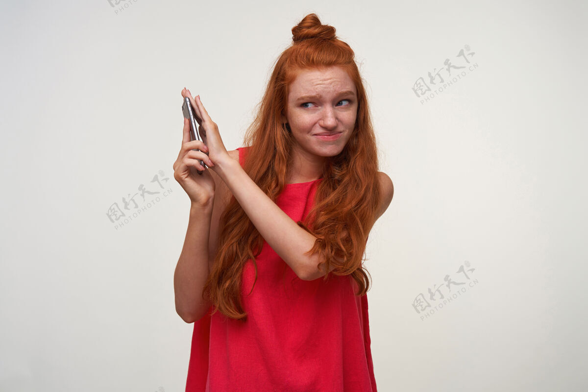 自然室内拍摄的年轻readhead女性头发打结 穿着休闲装在白色背景上摆姿势 用智能手机和手机进行不愉快的交谈 并遮盖手机以避免声音女士交谈休闲