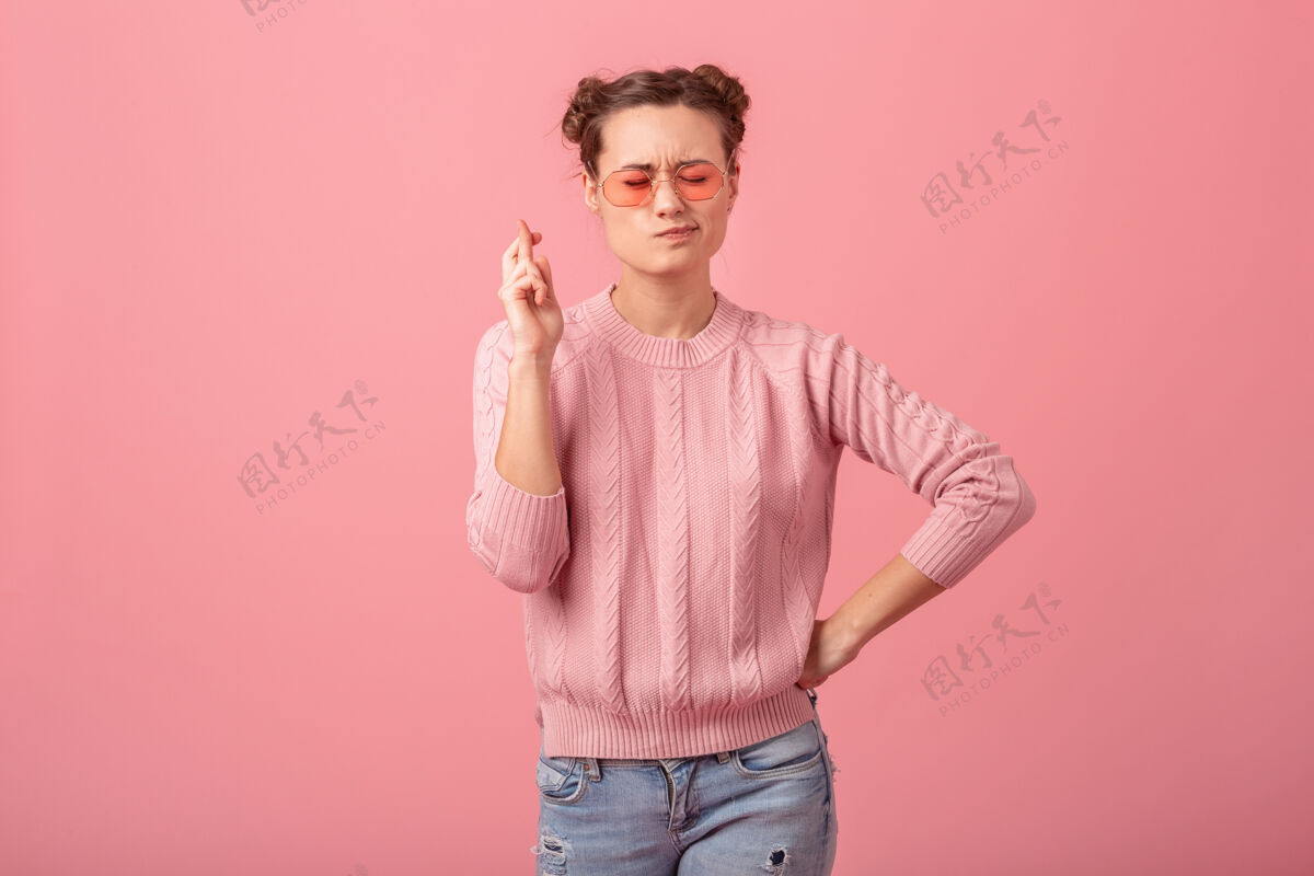 信仰年轻漂亮的女人做一个愿望交叉手指在粉红色的毛衣和太阳镜隔离在粉红色的工作室背景微笑粉色女孩
