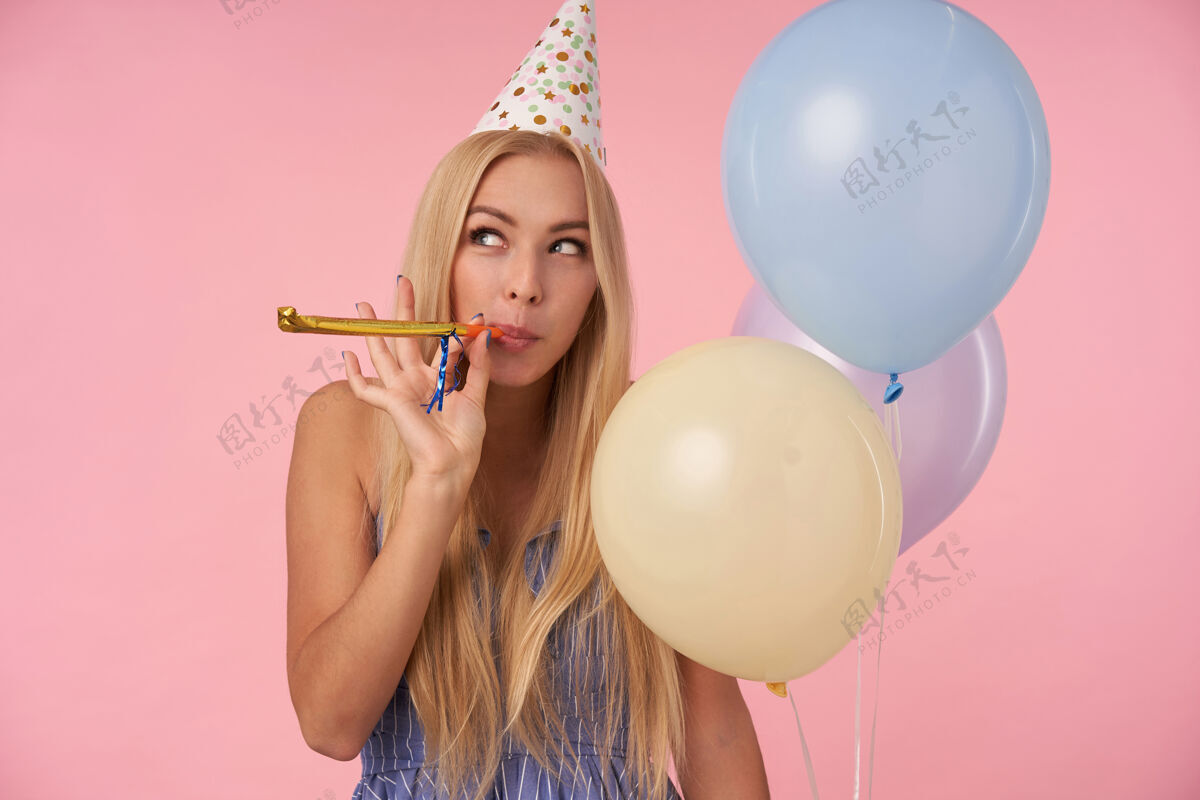 帽子美丽的金发长发女士嘴里叼着派对喇叭 积极地往一边看 在庆祝生日的时候摆出五颜六色的气球 站在粉色的背景上吹圆锥形金发