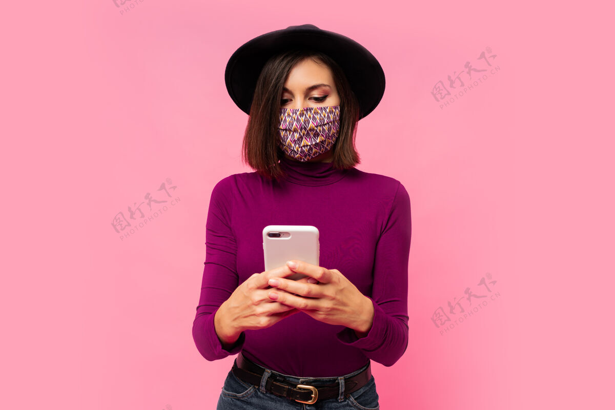 面具戴着防护面具和黑帽子的时髦女人用手机 站着使用粉色模型