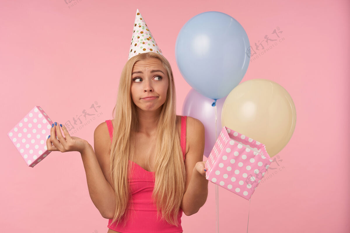 上衣困惑的年轻长发女性 随意的发型摆在粉色背景上 手里拿着礼物 庆祝节日 打开礼物 皱着嘴唇看着一边情感圆锥体撅嘴