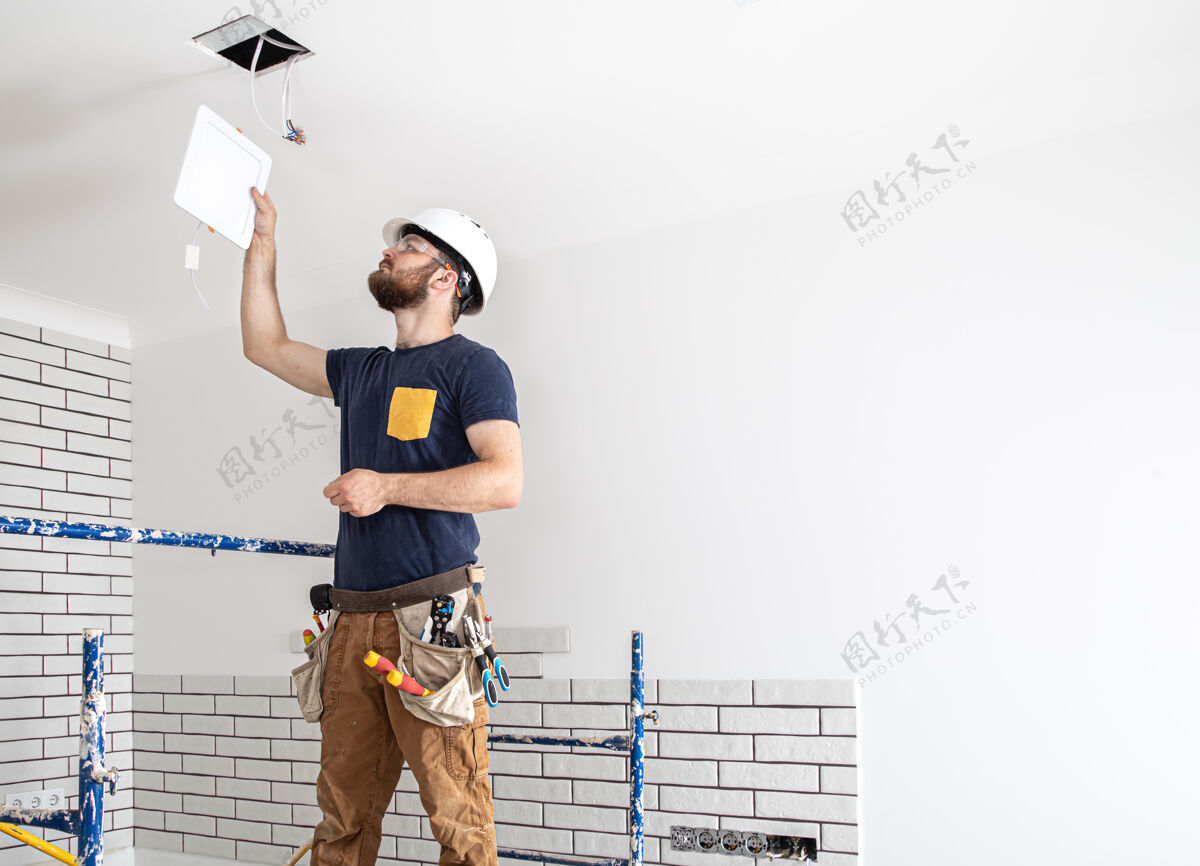 杂工电工建筑工人带着胡须 工人戴着白色安全帽 在高空安装灯具专业的工人穿着工作服 在维修现场的背景上带着电钻修理工安装复制