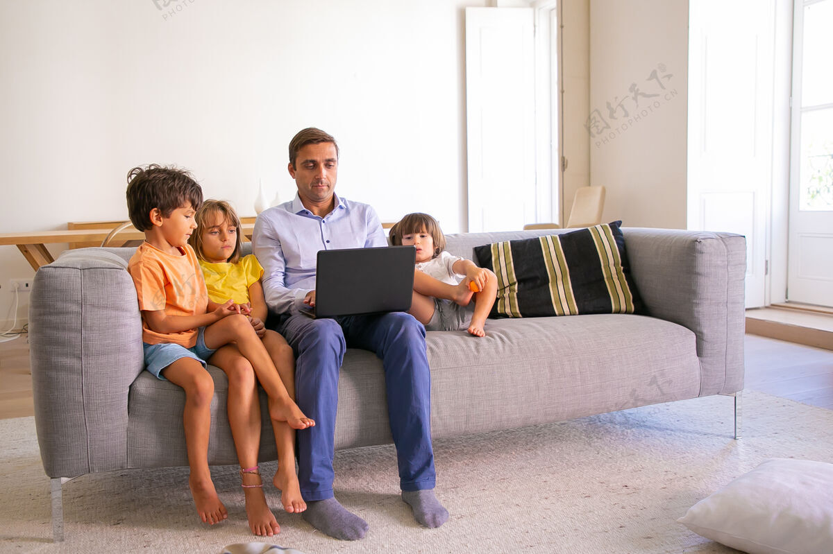 年轻专注的中年爸爸和孩子们坐在沙发上 用笔记本电脑打字白人爸爸和可爱的孩子在客厅里放松 看电影数字技术和父亲的概念沙发孩子朋友