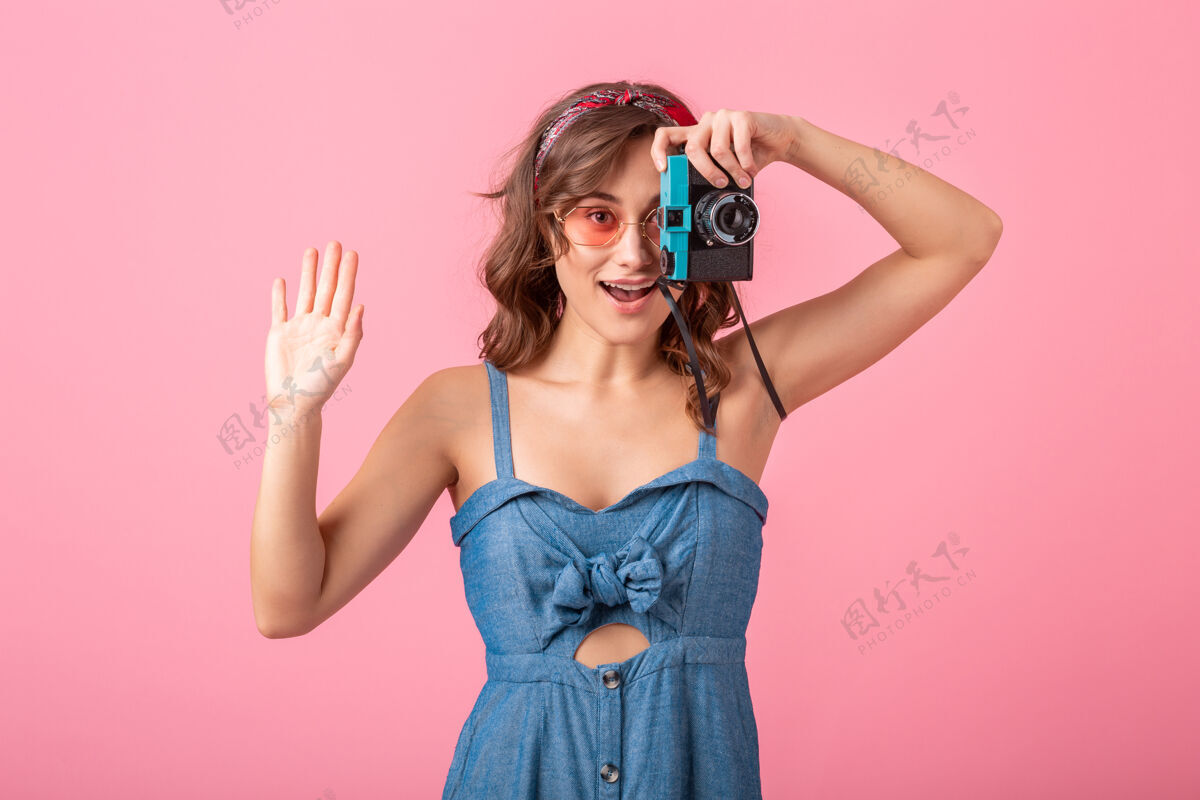 复古迷人的微笑女人在老式相机上拍照 手指向上 穿着牛仔裙 戴着粉色背景的太阳镜连衣裙女孩微笑