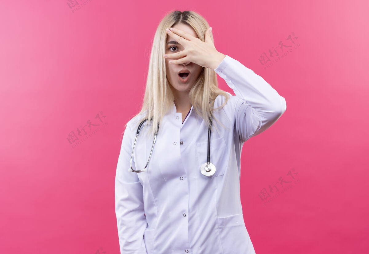 封面惊讶的医生年轻的金发女孩戴着听诊器在医疗长袍覆盖在孤立的粉红色背景的眼睛金发听诊器穿着