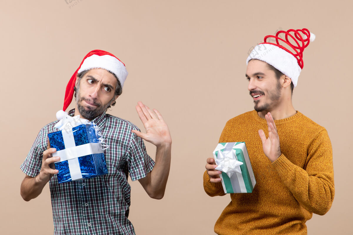 视图正面图两个家伙一个生气 一个幸福戴圣诞帽 拿着圣诞礼物在米色孤立的背景米色礼物帽子