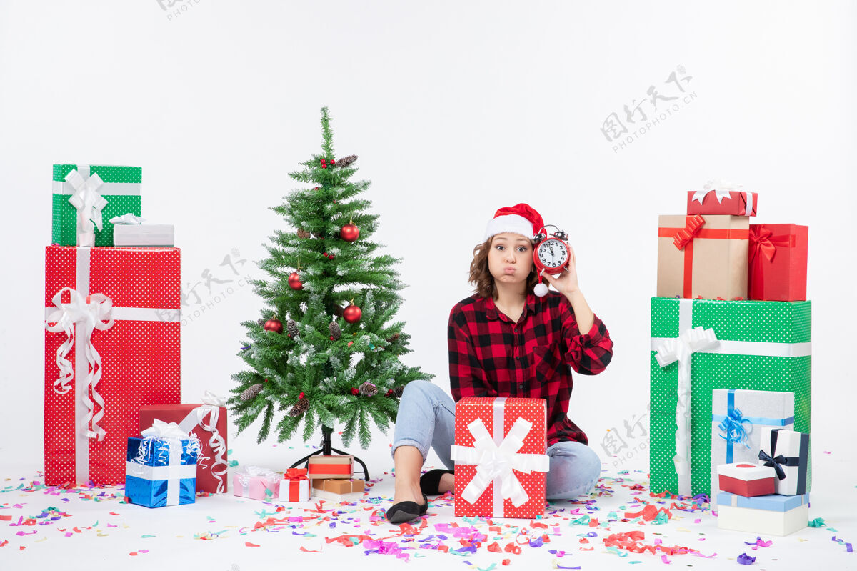 圣诞树前视图的年轻女子坐在圣诞礼物周围拿着白色墙上的时钟时钟女人节日