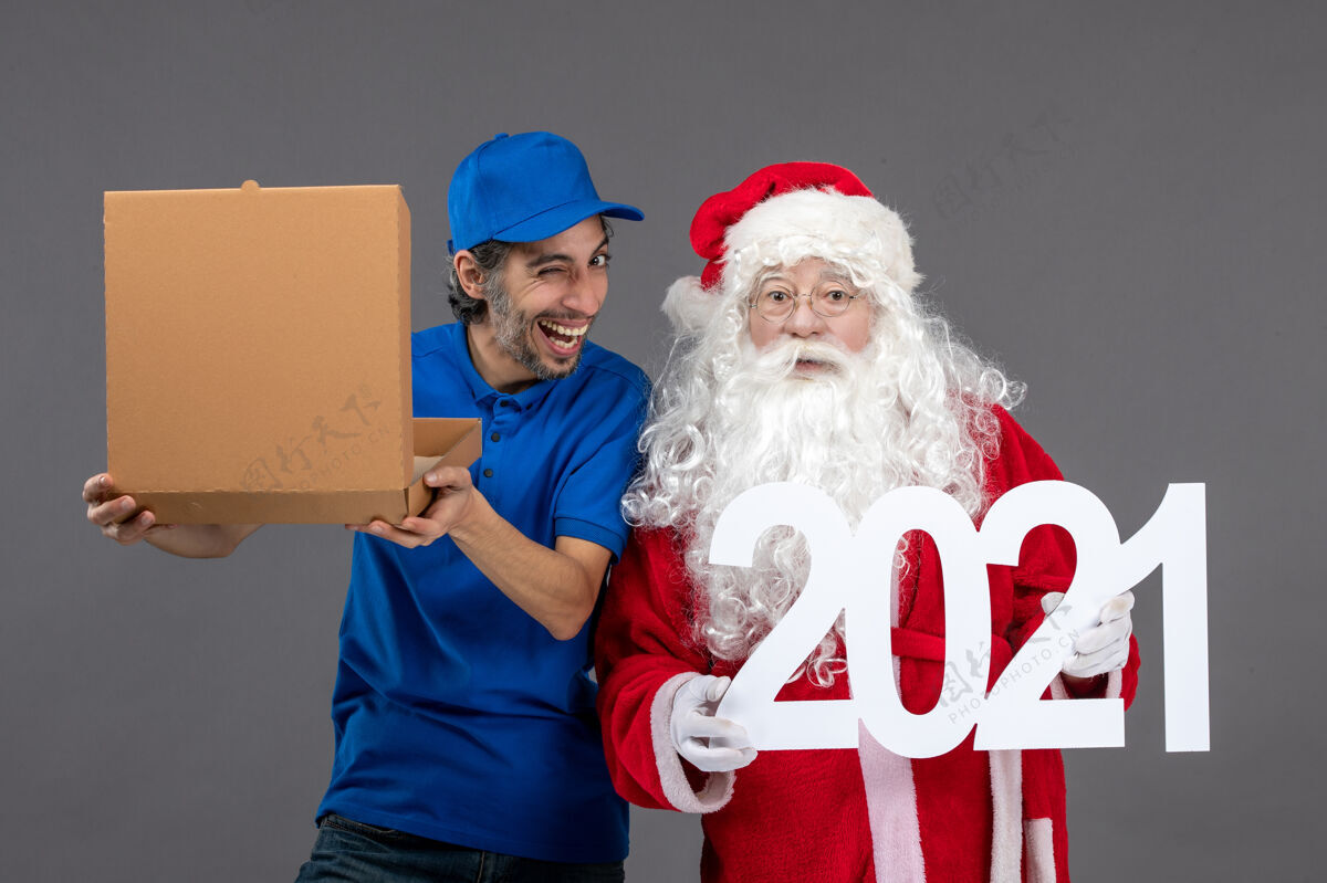 人圣诞老人的正面图 男信使拿着食物盒 在灰色的墙上写字前面快乐服装