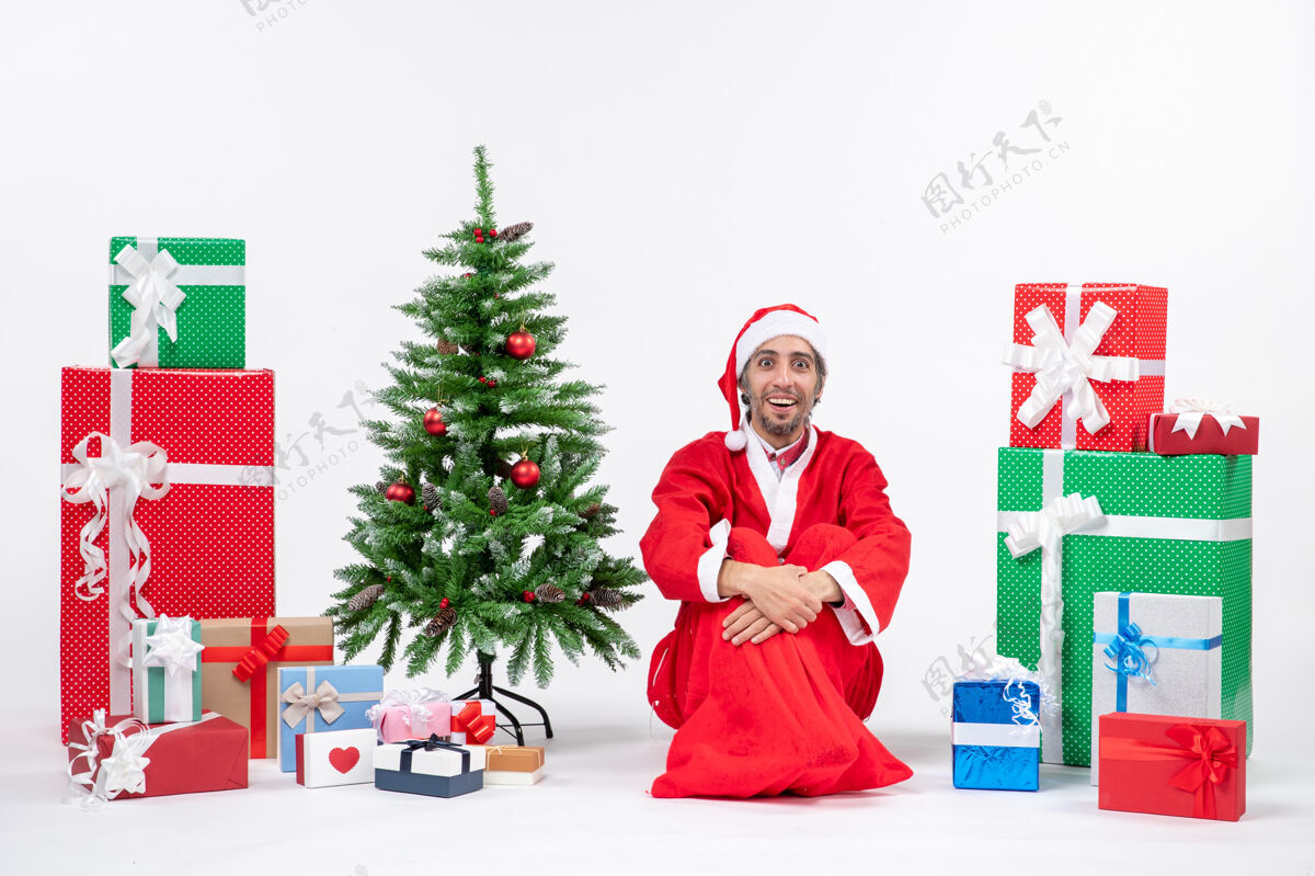 礼物微笑着惊讶的年轻人打扮成圣诞老人和礼物装饰圣诞树坐在地上的白色背景装饰微笑男人