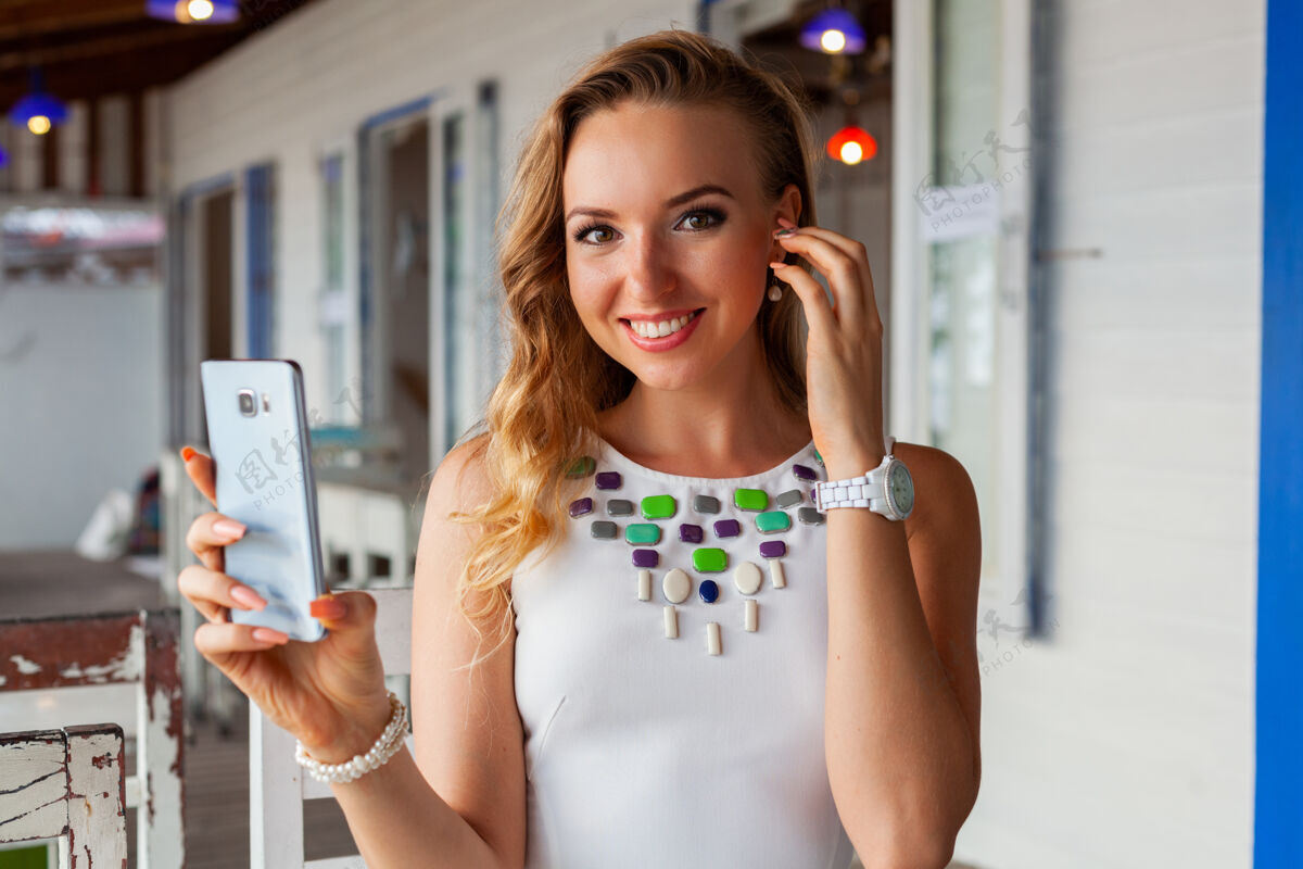 漂亮一位身着白色礼服的美女戴着墨镜在夏日咖啡馆用手机拍照手机户外放松