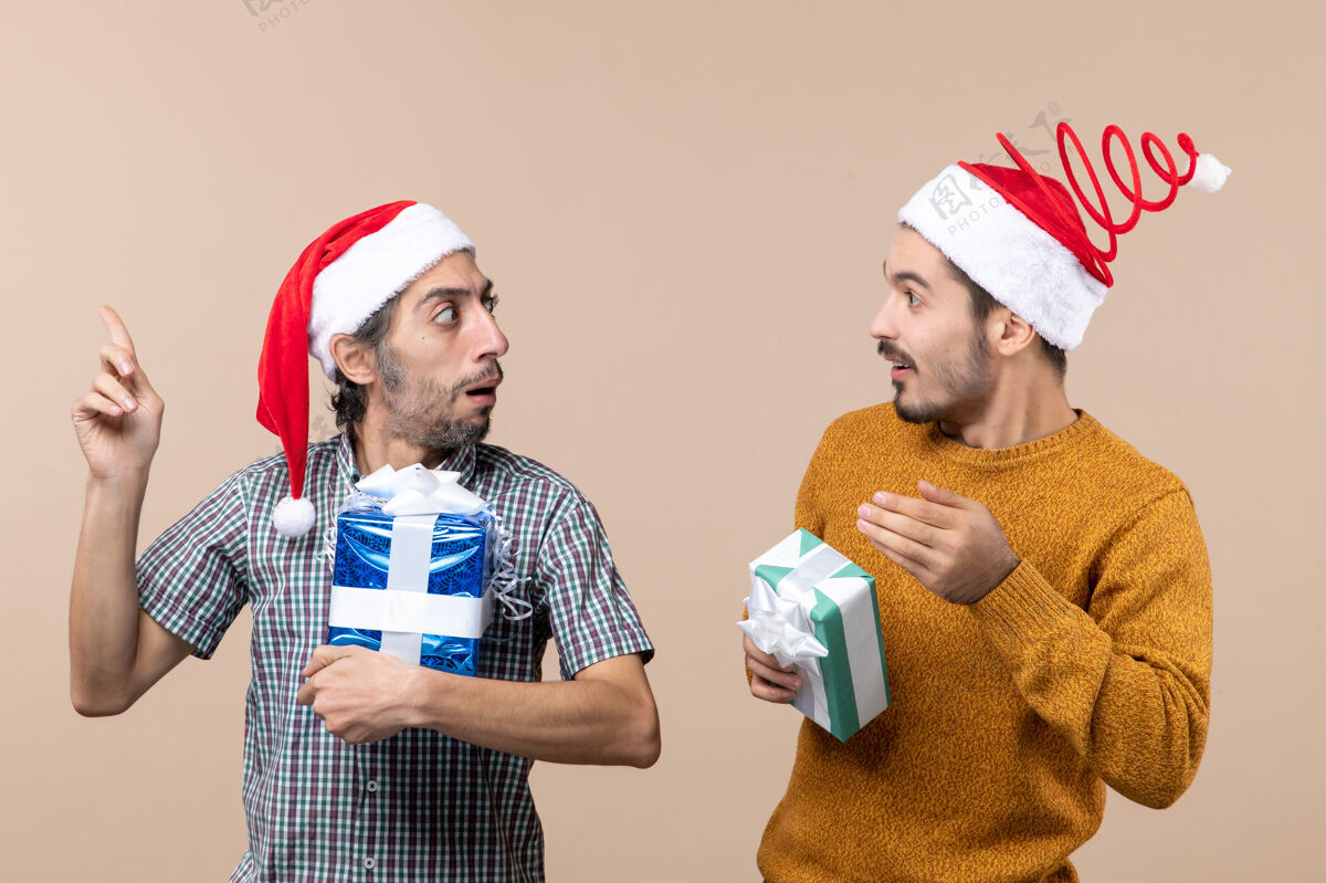 男性正面图两个有兴趣的家伙拿着圣诞礼物在米色的背景上相互展示着什么圣诞节米色伙计们
