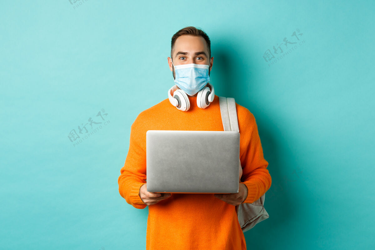 胡须年轻人戴着口罩用笔记本电脑 站在浅蓝绿色的墙上快乐男人现代