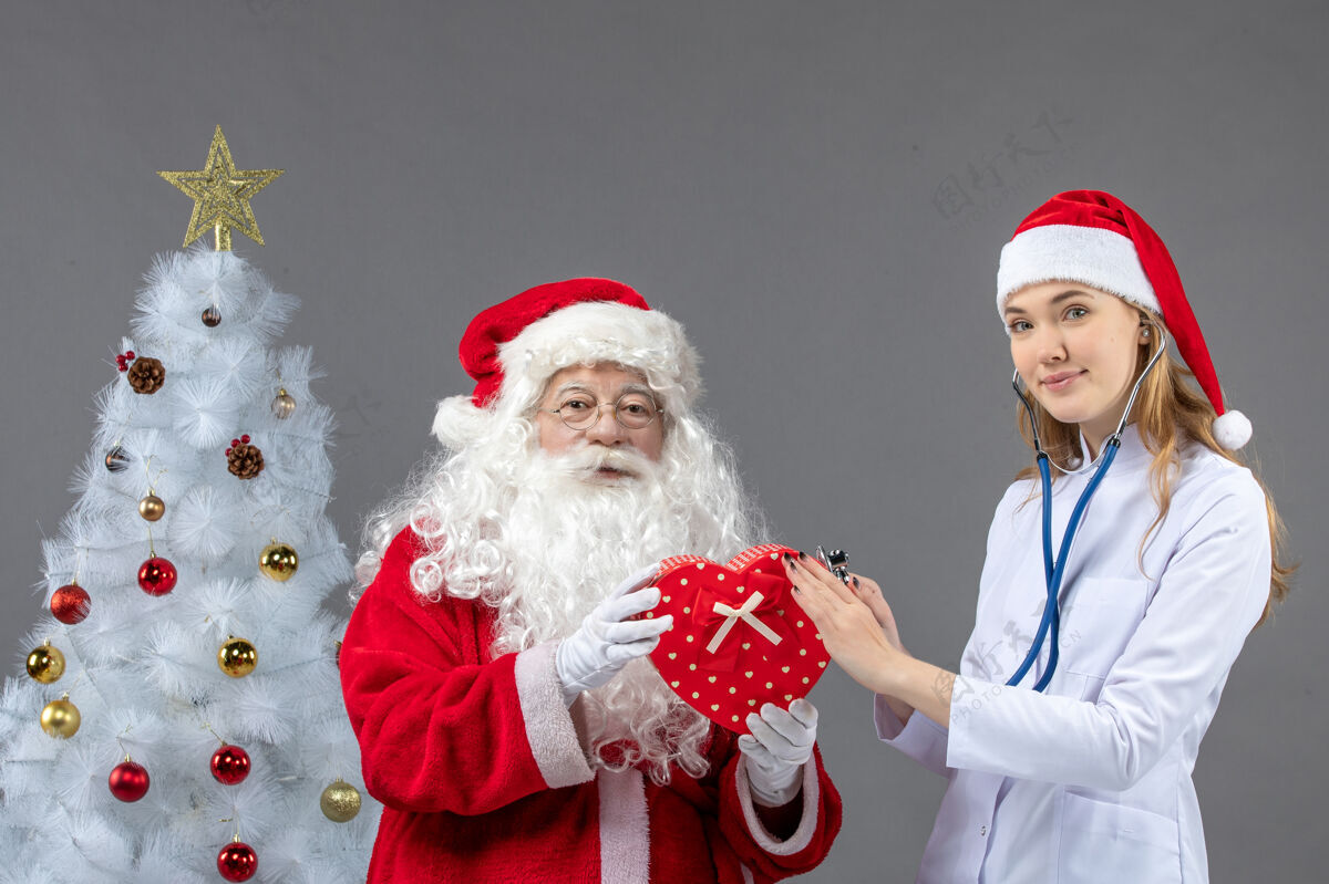 圣诞老人圣诞老人和女医生的前视图 女医生正在灰色墙上用听诊器观察礼物正面十二月医生