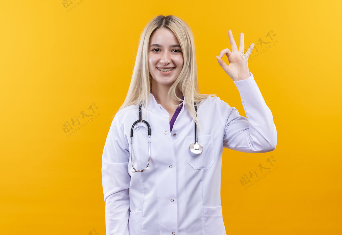 穿着微笑的医生年轻女孩穿着医用长袍戴着听诊器和牙套 在孤立的黄色背景上显示出良好的姿势手势牙科展示