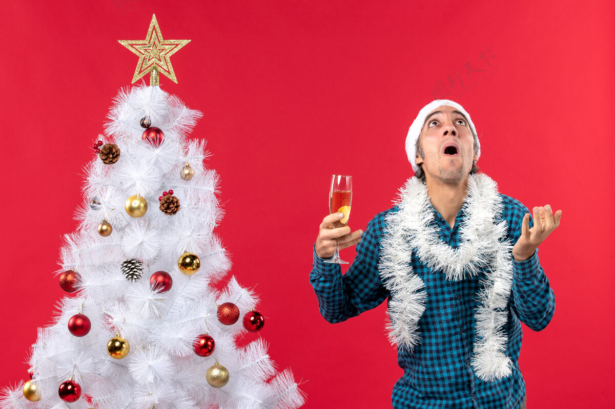 帽子圣诞节心情与情绪激动的年轻人与圣诞老人帽子在一个蓝色的衬衫剥离举行一杯葡萄酒在圣诞树附近抬头看年轻人圣诞老人家伙