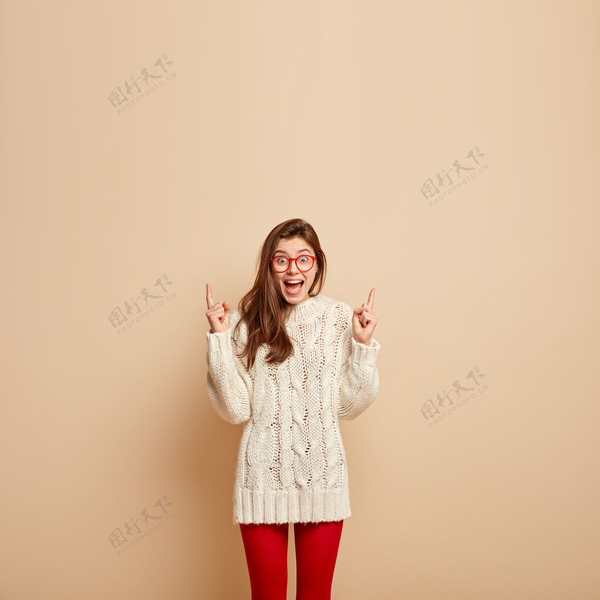 肖像照片中的正喜出望外的女性张大嘴巴 兴奋地尖叫 在自由空间向上指 穿着白色套头衫 戴着透明眼镜 隔离在米色的墙上手指站立手势