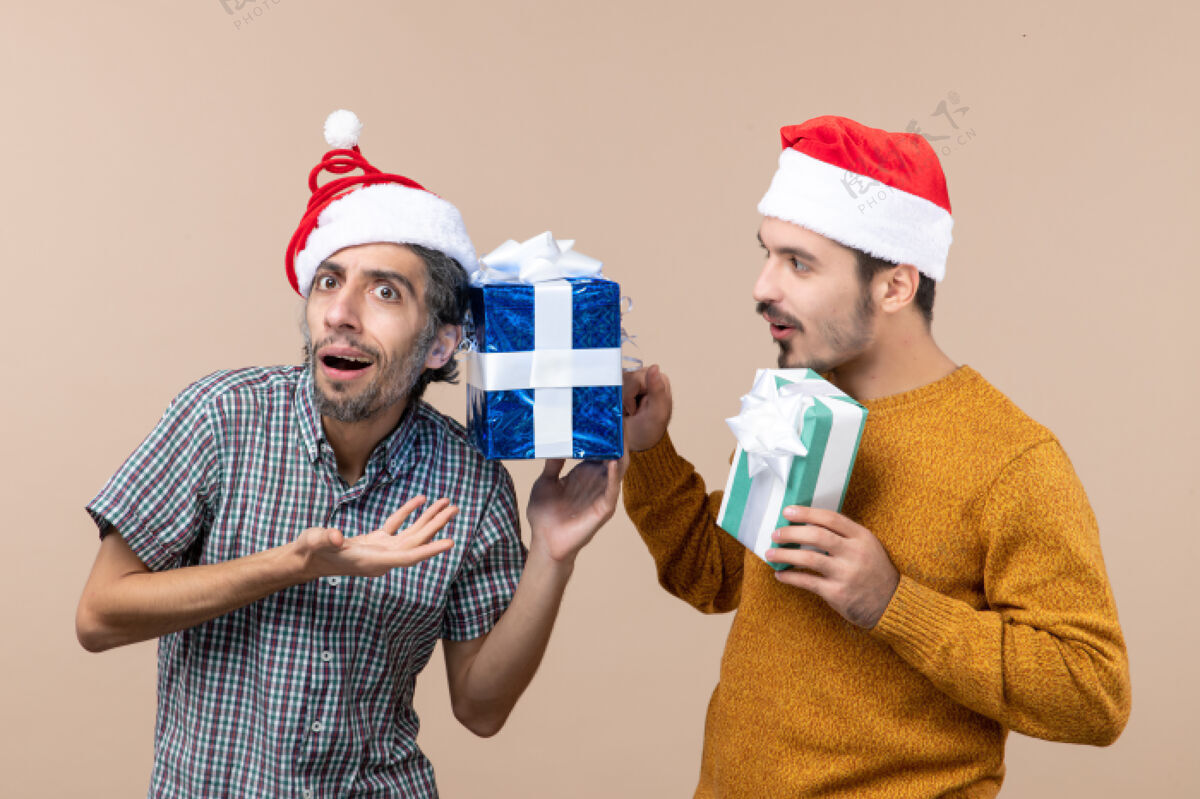 前面前视图两个困惑的家伙戴着圣诞帽和检查他们的圣诞礼物米色孤立的背景伙计们帽子男人