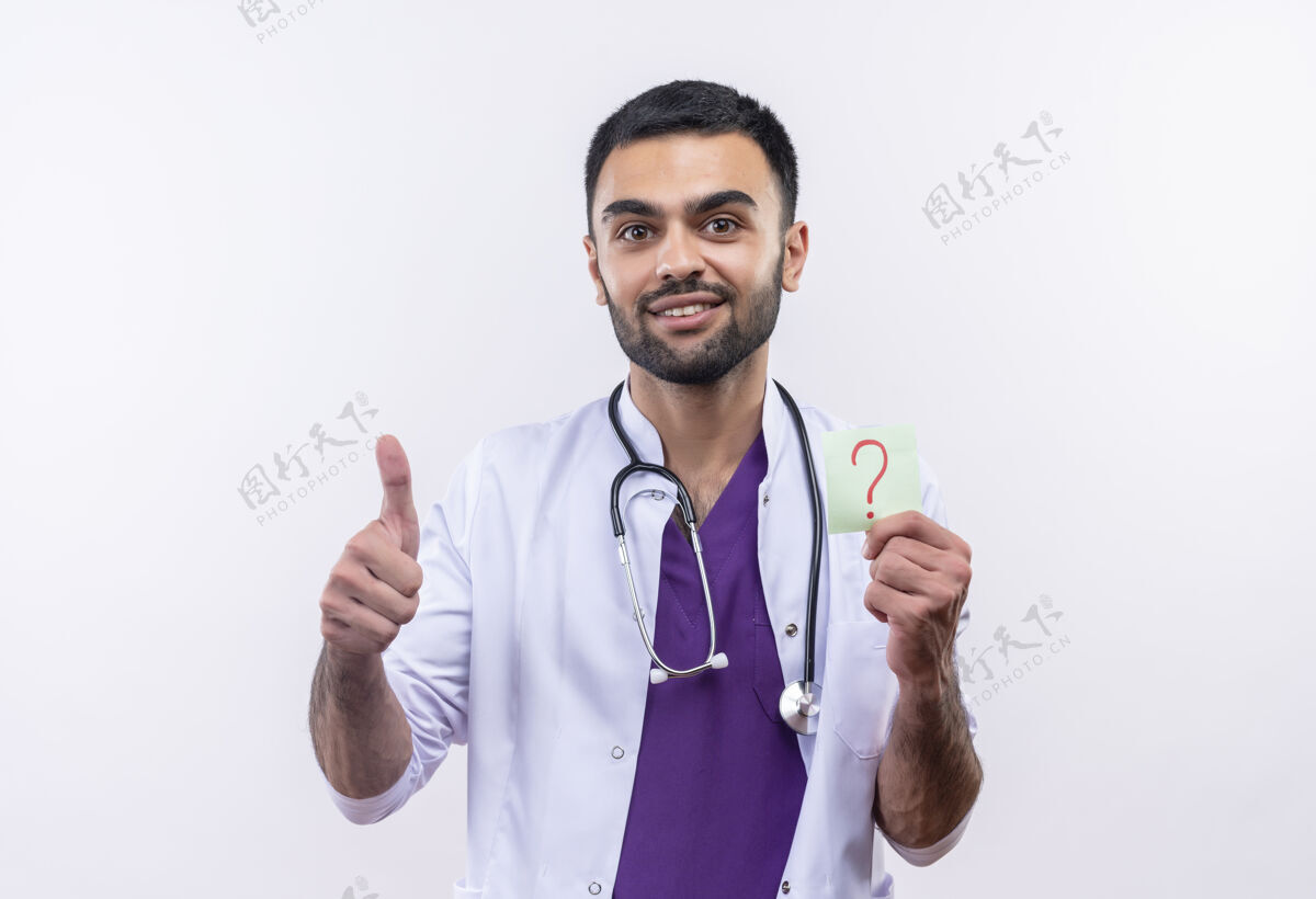 听诊器微笑着的年轻男医生穿着听诊器医用长袍拿着纸问号在孤立的白色背景上竖起大拇指白色问号男性