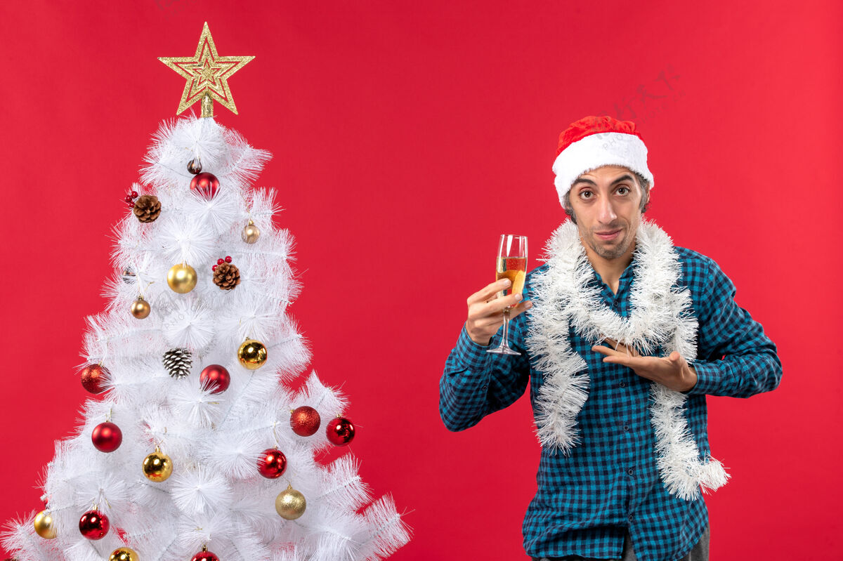 前夜圣诞心情自信的年轻人 戴着圣诞老人帽 穿着蓝色条纹衬衫 在圣诞树旁举起一杯葡萄酒庆祝圣诞树玻璃