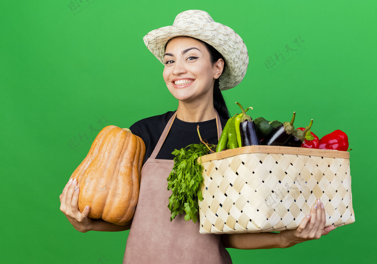 脸年轻漂亮的女园丁围着围裙 戴着帽子 手里拿着装满蔬菜和南瓜的箱子看着镜头 满脸笑容站着女人满满的