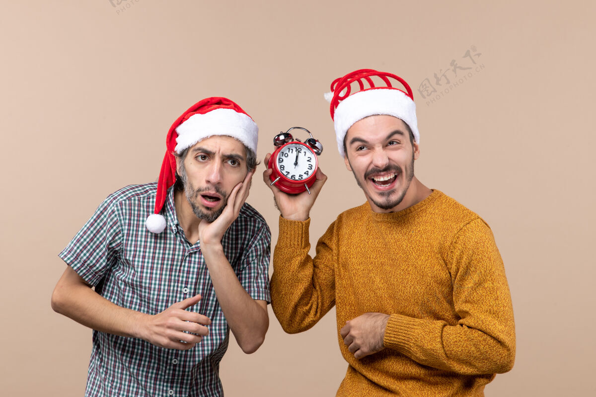 男人前视图两个男人一个困惑 另一个拿着闹钟 而笑着在米色孤立的背景时钟迷惑帽子脸