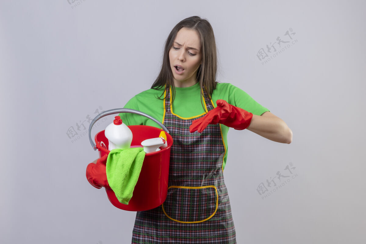 清洁悲伤的清洁年轻女孩穿着制服 戴着红手套 指着她手上的清洁工具 背景是孤立的白色红白戴