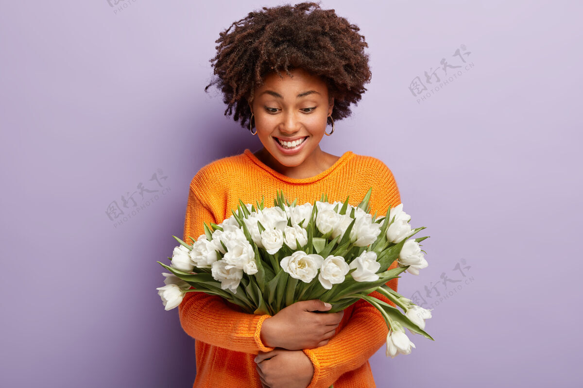 满意温柔积极的非洲裔美国女士拥抱着温柔的白色花朵 微笑着看着郁金香 穿着橙色的套头衫非洲女性种族