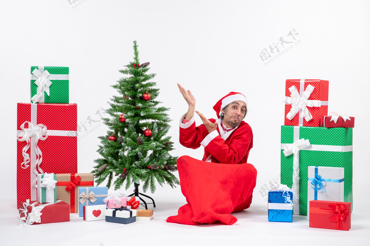 成人惊讶的年轻人打扮成圣诞老人与礼物和装饰圣诞树坐在地上指着白色背景的右边的东西礼物礼物打扮