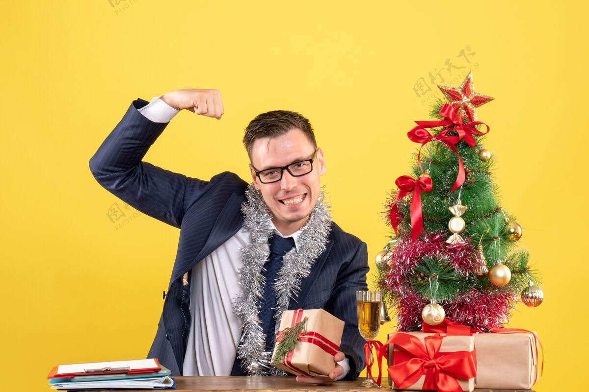 男人年轻人展示肌肉坐在圣诞树附近的桌子上 并在黄色的礼物男年轻人礼物