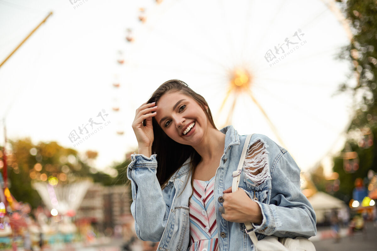 城市年轻漂亮 快乐的黑发女性 穿着时髦的牛仔裤外套 站在游乐场的摩天轮旁 开心地看着 拉直头发棕色衣服姿势