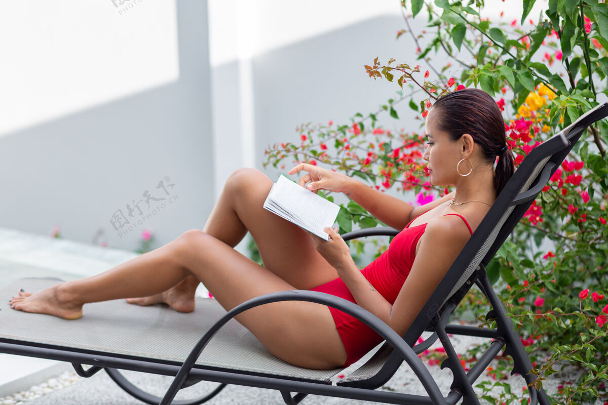 平静穿着泳衣的亚洲女人在日光浴床上的泳池边放松 在五颜六色的花树旁的别墅里看书热带休闲时尚
