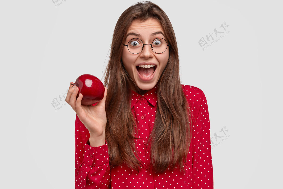 维生素快乐的黑发女人张大嘴巴 有快乐的表情 戴着圆眼镜 红色圆点衬衫 捧着新鲜的苹果饮食苹果黑发