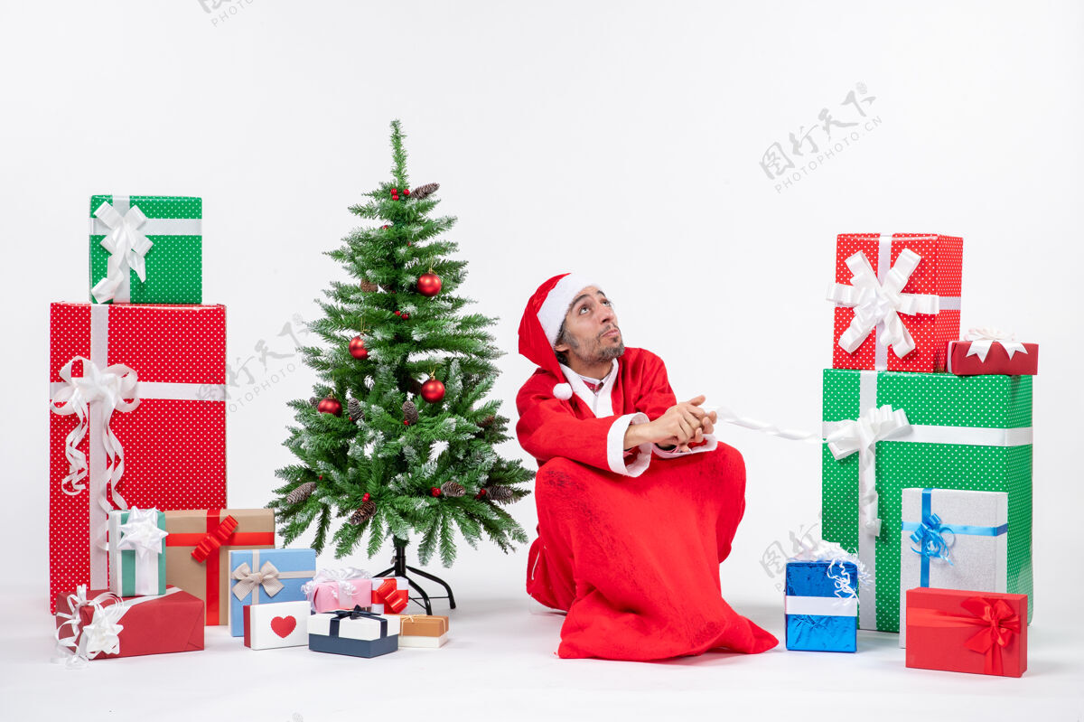 地面专注的年轻人打扮成圣诞老人 拿着礼物和装饰好的圣诞树坐在白色背景的地上礼物年轻人装饰