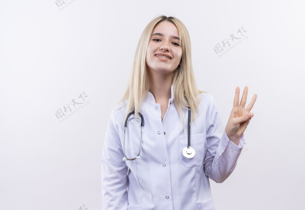 医生微笑的医生 年轻的金发女孩 戴着听诊器 穿着医用长袍 戴着牙套 在孤立的白色背景上显示三个女孩三个长袍
