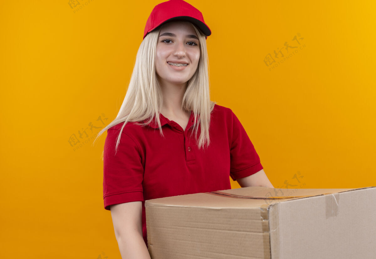 牙科微笑的小女孩穿着红色t恤 戴着帽子 戴着牙套 在孤立的橙色背景下拿着盒子抱着送货微笑