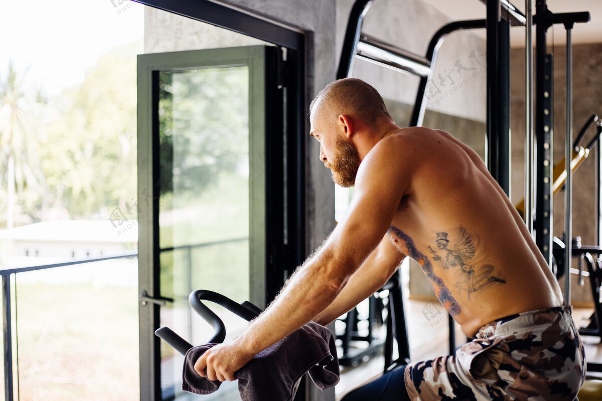 运动装纹身 肌肉发达 胡须浓密的男人骑着自行车在体育馆里锻炼有氧运动 靠近大窗户 外面可以看到树训练亚洲人健康