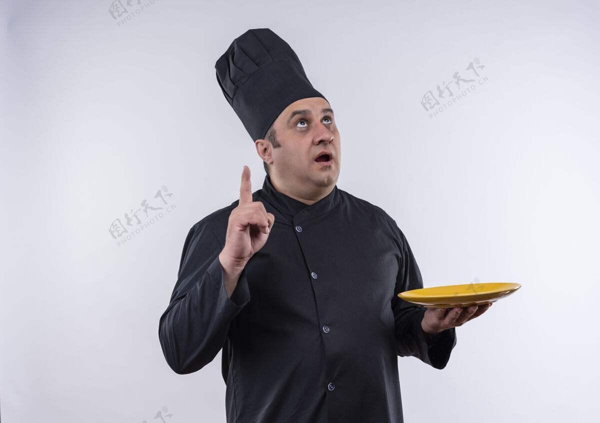 惊讶抬头一看 惊讶的中年男厨师穿着厨师制服 拿着盘子指着孤立的白墙上的手指厨师制服中年
