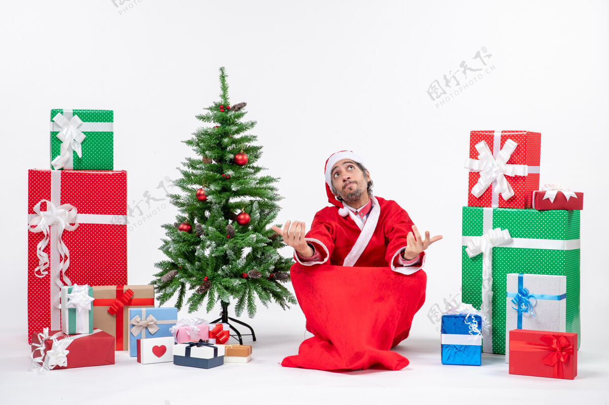 年轻人惊讶激动困惑的年轻人打扮成圣诞老人带着礼物和装饰圣诞树坐在白色背景的地上人礼物圣诞