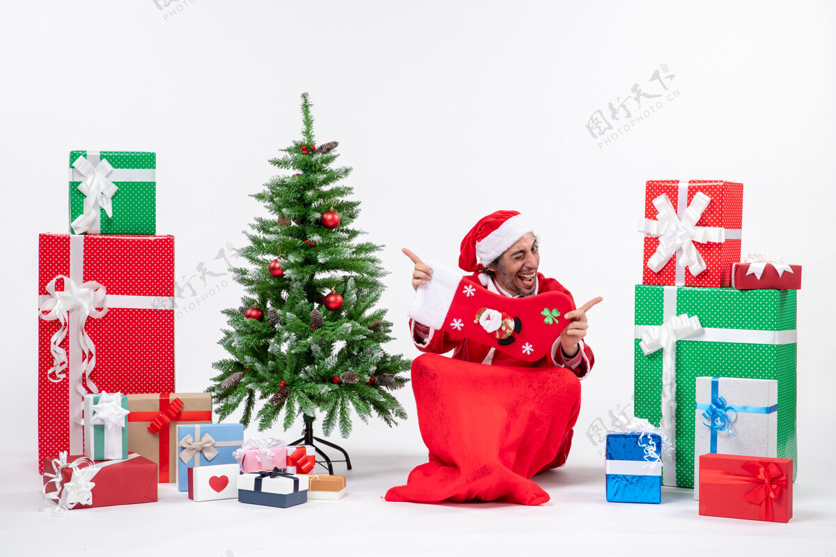 地面新年气氛与有趣的圣诞老人坐在地上 并显示圣诞袜子附近的礼物和装饰圣诞树上的白色背景圣诞树圣诞装饰