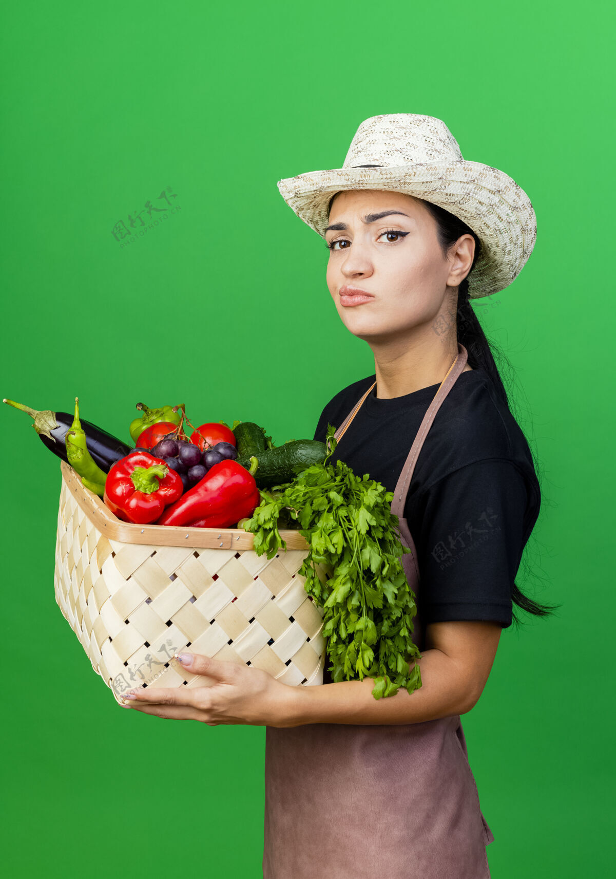 绿色年轻漂亮的女园丁 围着围裙 戴着帽子 手里拿着装满蔬菜的篮子 带着怀疑的表情园丁帽子女人