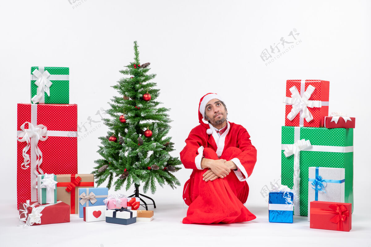 年轻人困惑的年轻人打扮成圣诞老人与礼物和装饰圣诞树坐在地上的白色背景微笑年轻人坐着