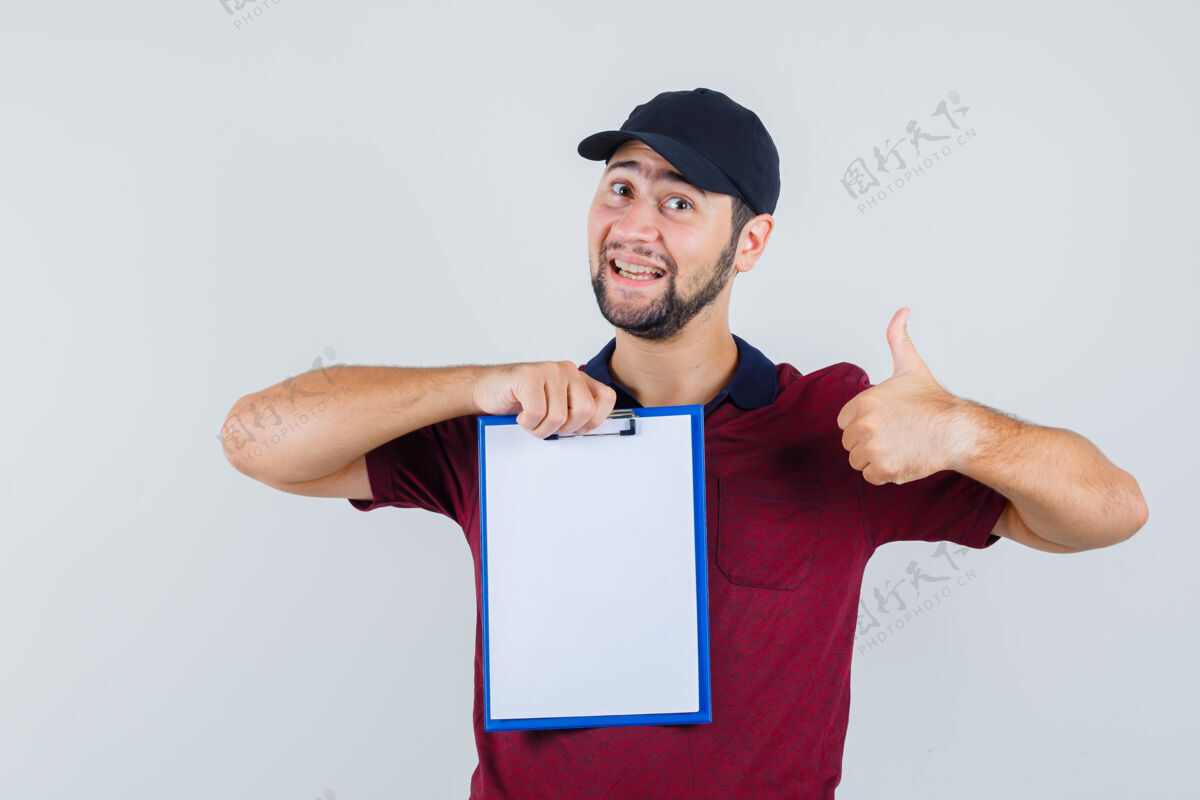 休闲年轻的男性在展示笔记本的时候竖起大拇指 穿着红色t恤 戴着黑色帽子 看上去很开心正面图帅气站立小伙子