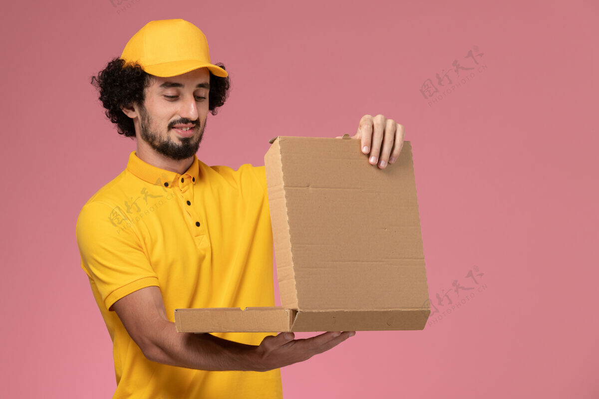 食物正面图：身穿黄色制服的男快递员拿着食物盒站在浅粉色的墙上视图持有披萨