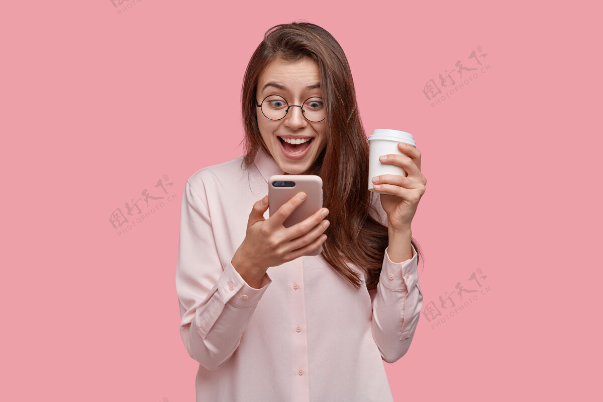 手机印象深刻的美女喜出望外 兴奋地看短信 穿得正式 端着外卖咖啡 隔着粉色的工作室墙人印象深刻眼镜小玩意
