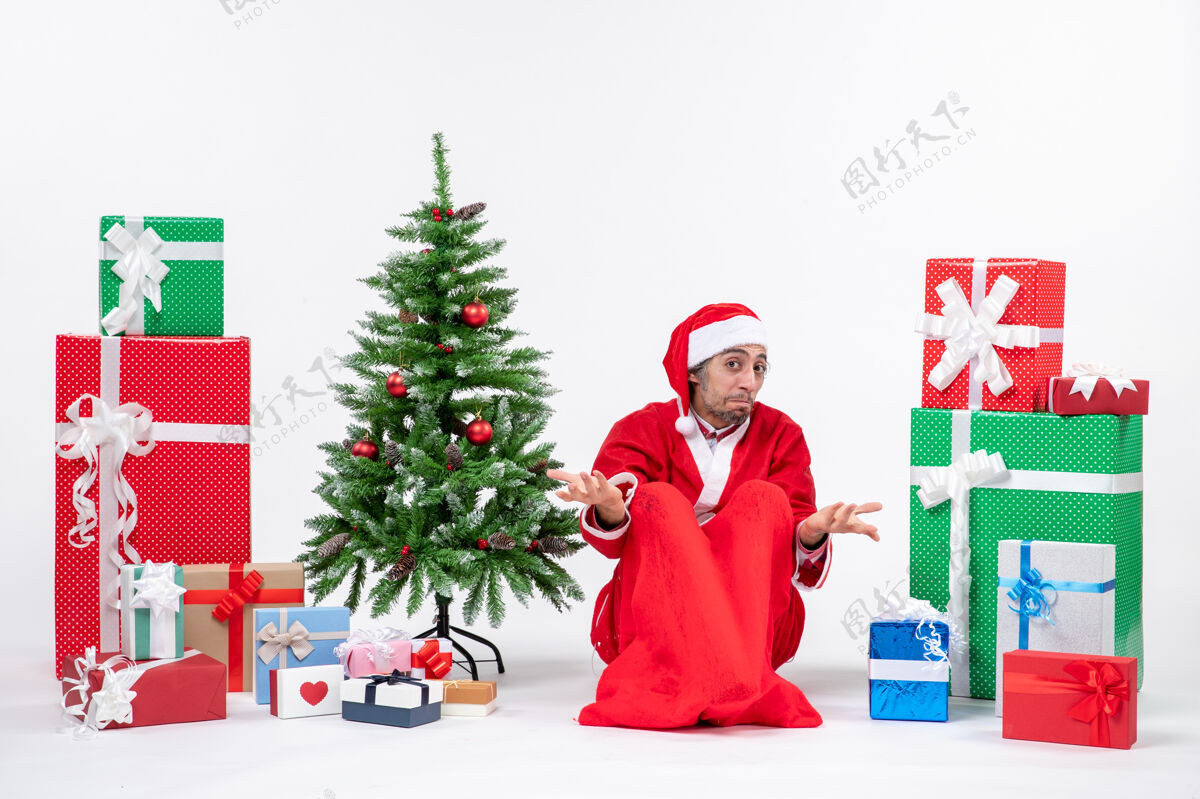 坐着困惑的年轻人打扮成圣诞老人 拿着礼物和装饰过的圣诞树坐在地上的白色背景上圣诞老人圣诞老人年轻人