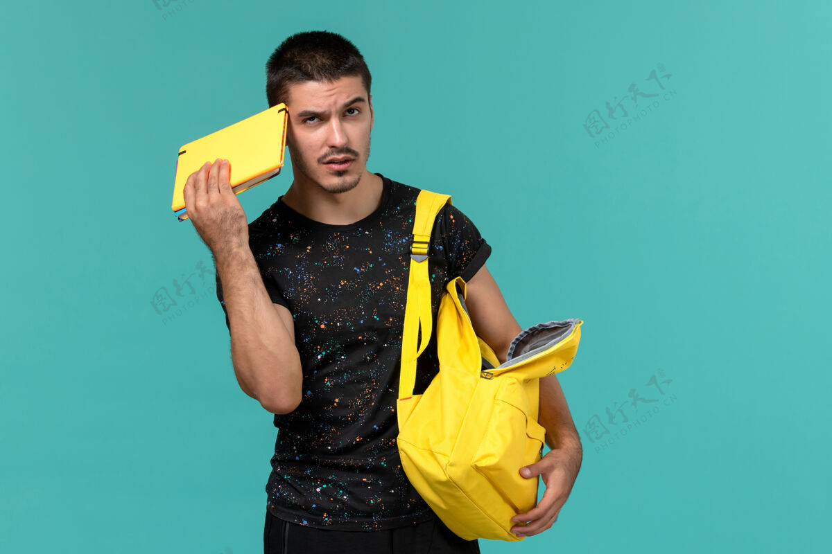 可爱身穿深色t恤黄色背包的男学生在浅蓝色墙上思考的正面视图大学快乐男性